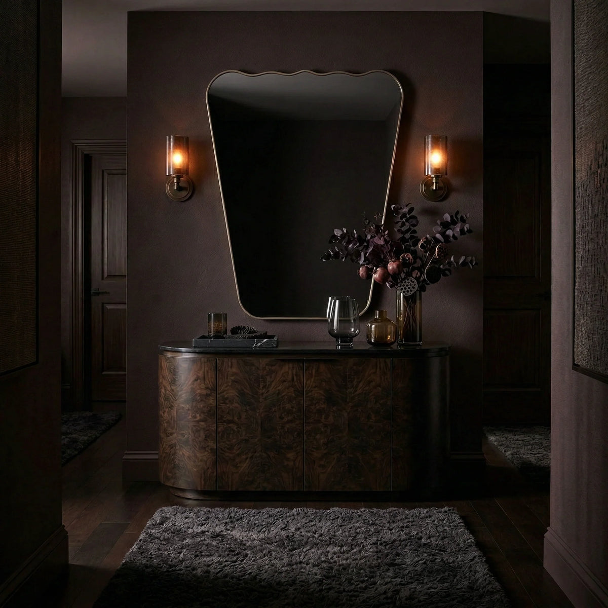 38 Wavy Mirror - Antiqued Brass by Ambella Home styled above a curved dark wood credenza in a moody luxury entryway, featuring an organic tapered silhouette, softly waved top edge, and warm antiqued brass frame