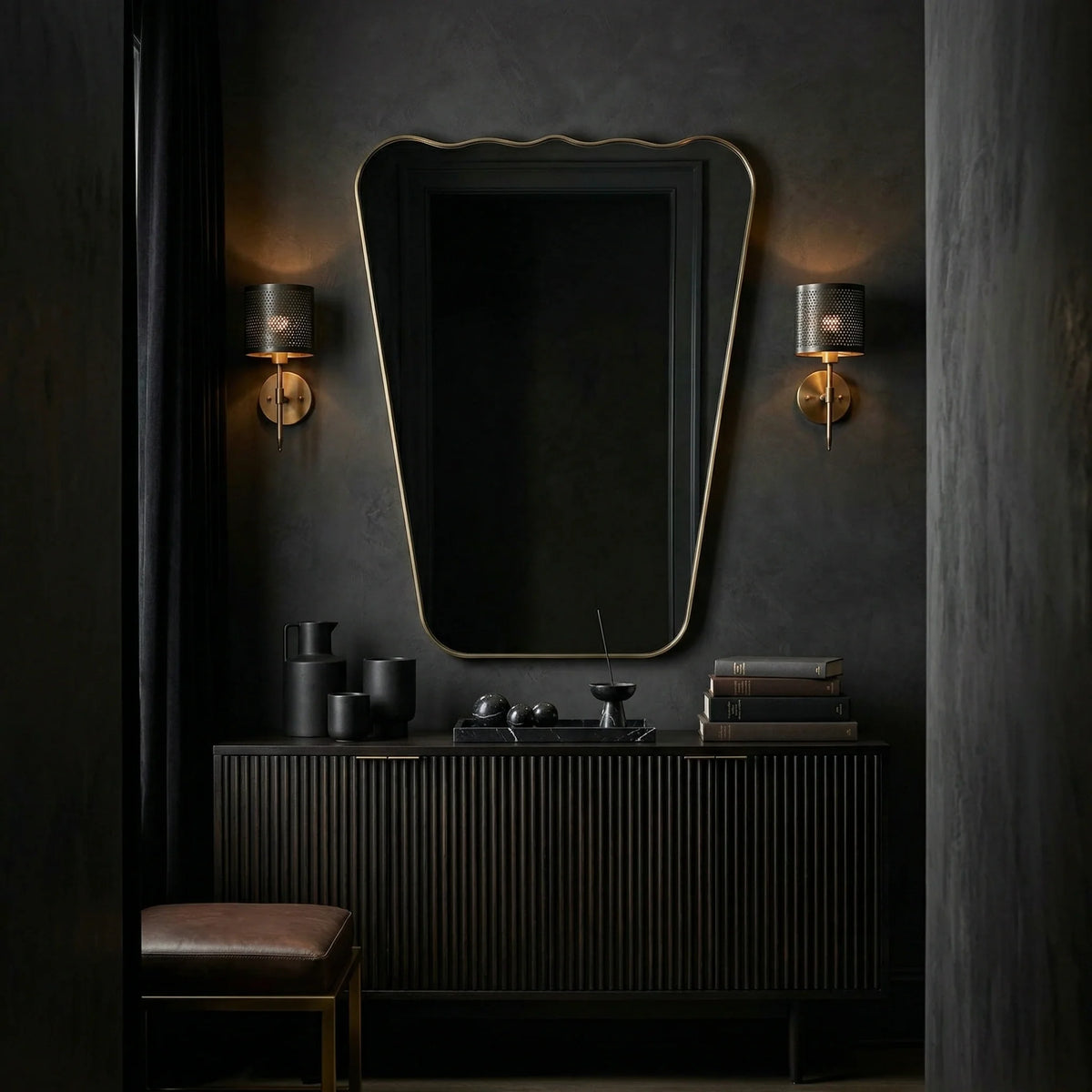 Ambella Home 38 Wavy Mirror in antiqued brass displayed over a fluted black credenza in a dramatic dark hallway, showcasing a sculptural tapered profile with a gently undulating top and refined metallic frame