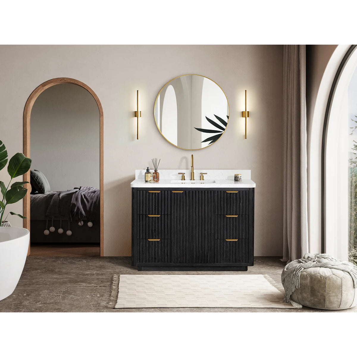 Wide view of the vanity below a mirror, arched openings behind and a pouf at the right