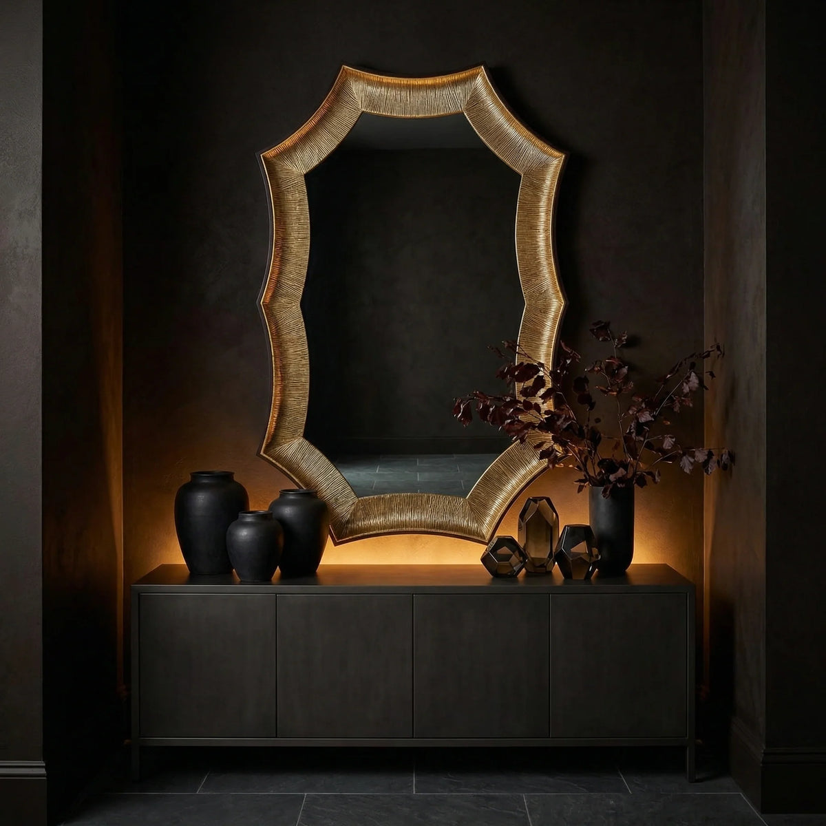 Ambella Home Helios Mirror Large, bold sculptural accent mirror with textured gold frame, centered over a dark credenza in an elegant black hallway with contemporary styling
