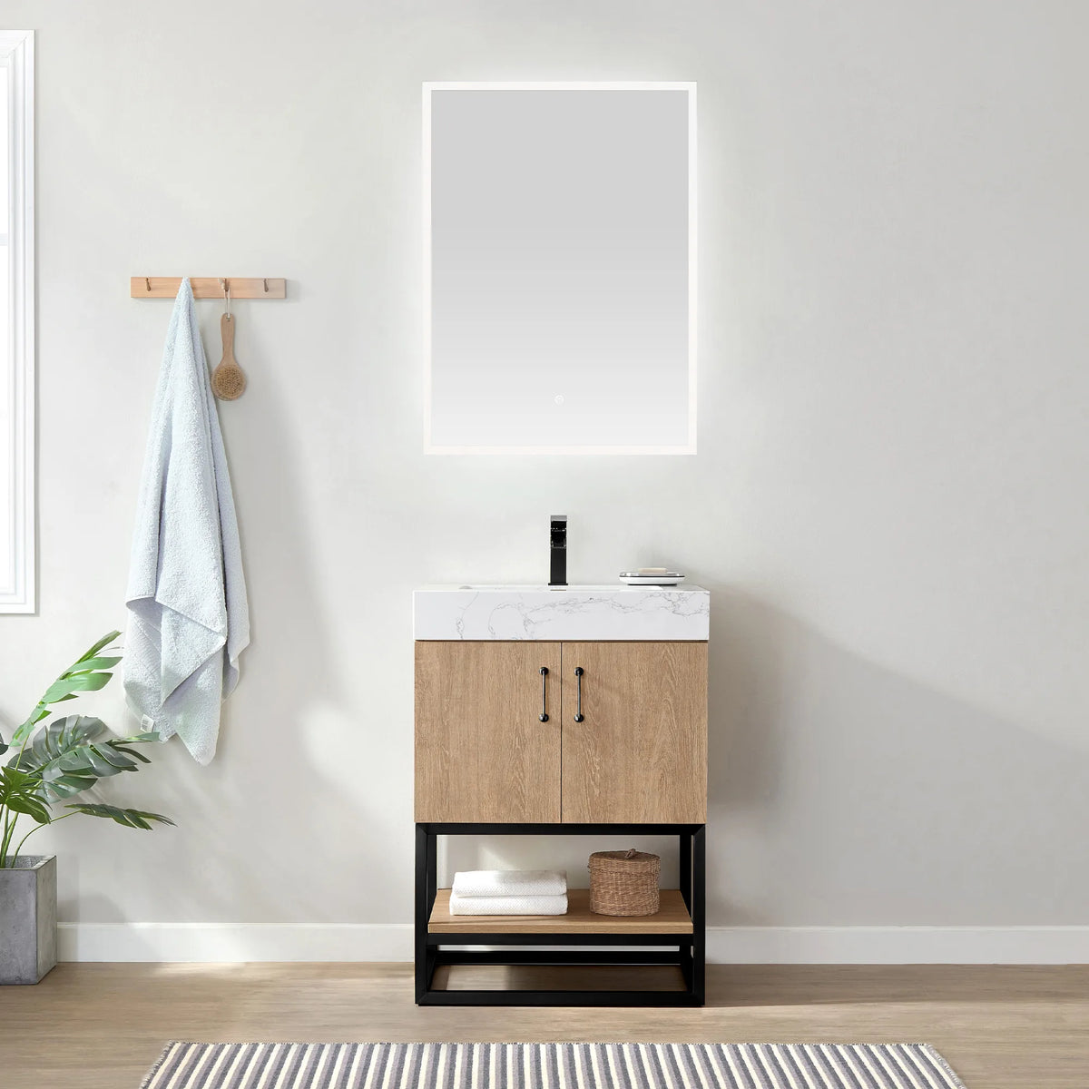Room view with the lit mirror above the vanity, towel on a peg rail, potted plant and striped rug