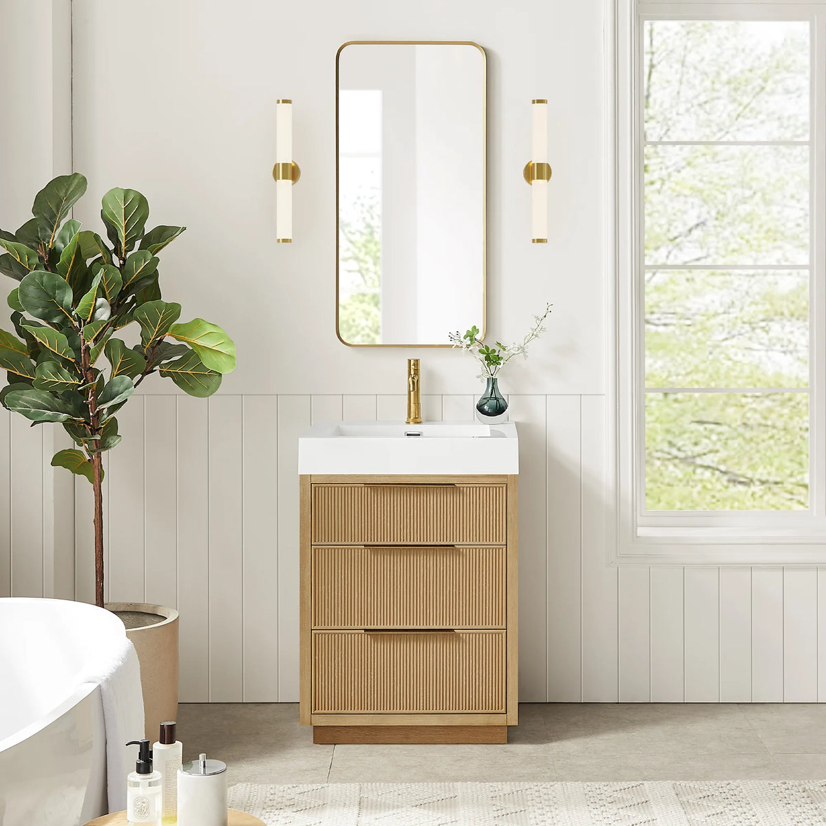 Window-lit bathroom setting for the 24-inch vanity, with gold-framed mirror and sconces; a bathtub is in view