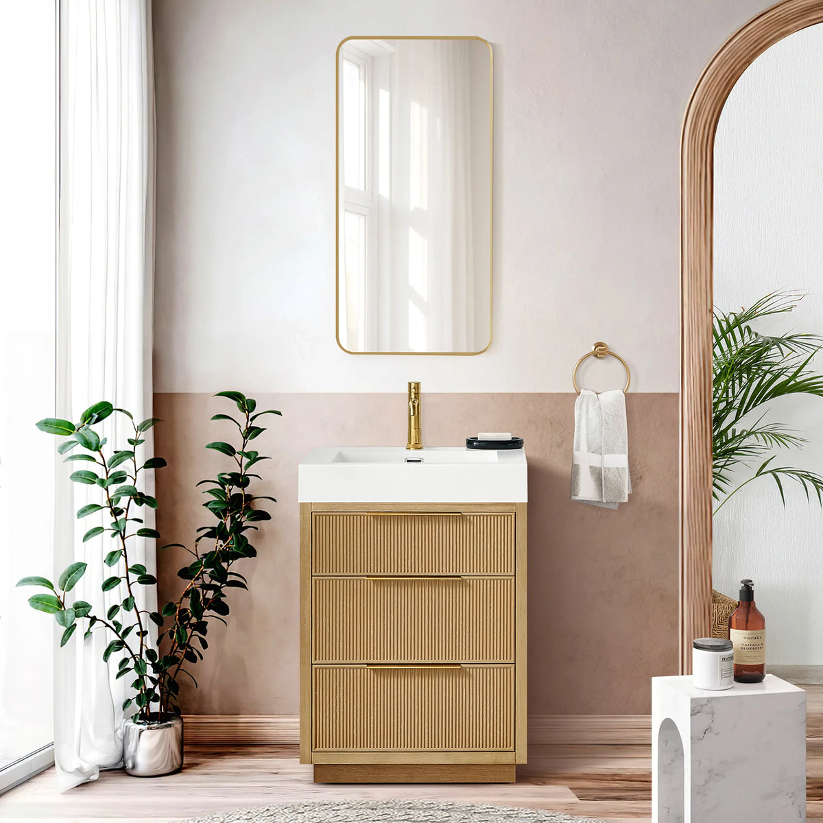Lifestyle shot of the 24-inch single vanity against a rose wall, with a gold-framed mirror and a towel ring; plants in view